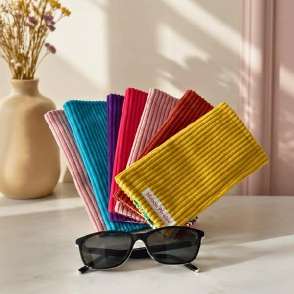 Éventail d'étuis à lunettes en velours côtelé de différentes couleurs (rose, bleu, violet, rouge, terracotta, jaune) avec des lunettes noires au premier plan.