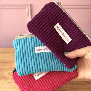 Maison Pivoine Une main tient deux pochettes à fermeture éclair en tissu côtelé, l'une bleue et l'autre violette, portant chacune l'étiquette de Maison Pivoine. Le Lotus rouge de Porte-monnaie est partiellement visible en dessous. Fait main en Bretagne