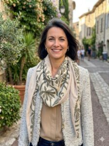 foulard-femme-printanier Portrait d'une femme souriante portant le chèche réversible Maison Pivoine sur une veste beige dans une ruelle ensoleillée.