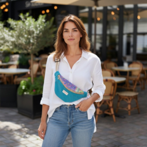 Maison Pivoine Une femme en chemise blanche et jeans se tient devant un café, portant le Sac banane velours "Musa" en bleu et regardant la caméra. Fait main en Bretagne