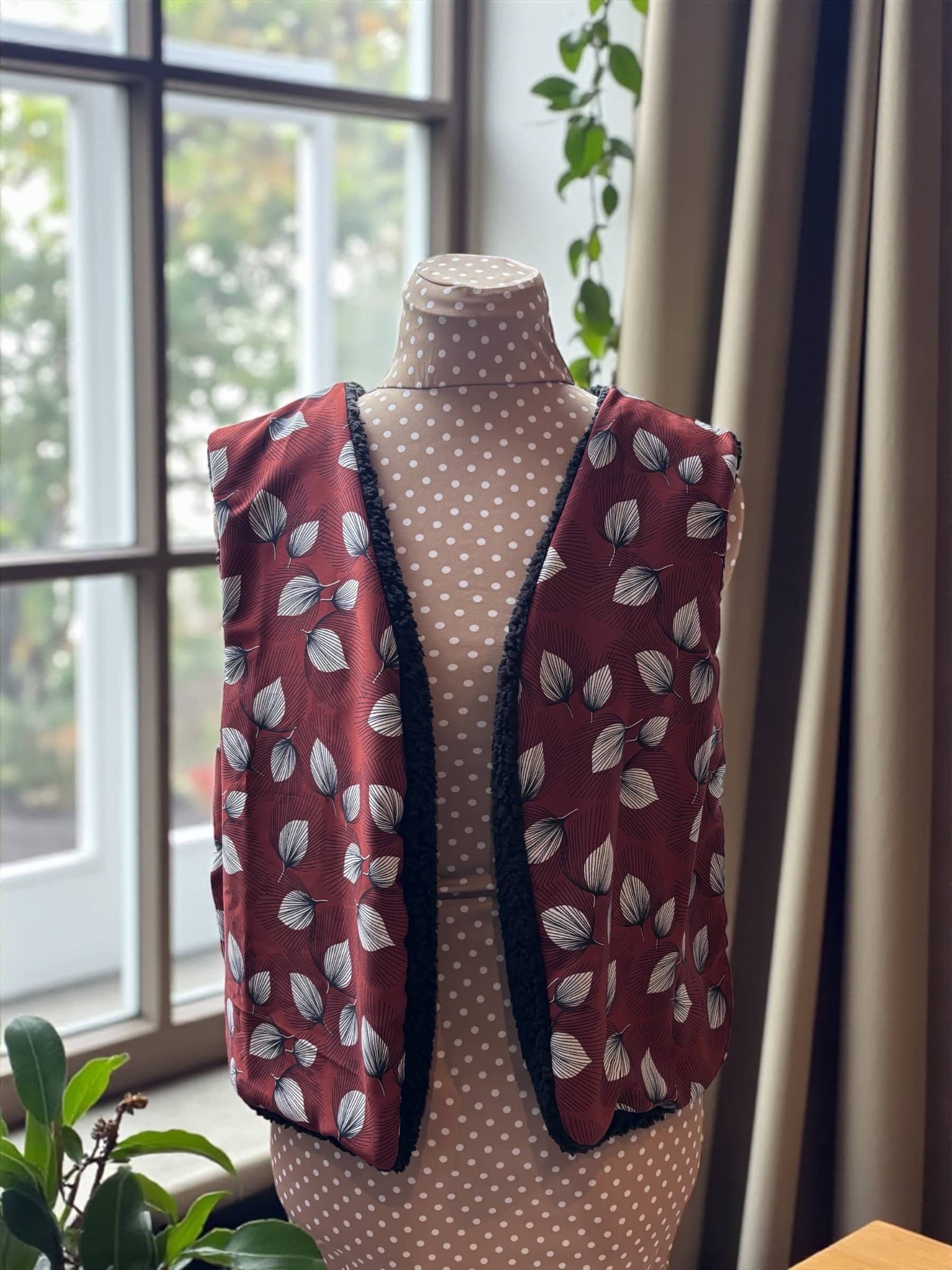 Gilet de berger sans manches réversible présenté sur mannequin, côté viscose chocolat à motif feuilles, avec bordures en sherpa noir, près d’une fenêtre.