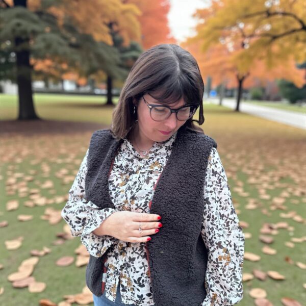 Femme portant un gilet de berger sans manches en sherpa noir par-dessus une blouse fleurie, en extérieur dans un parc avec des feuilles d’automne au sol, regard baissé.