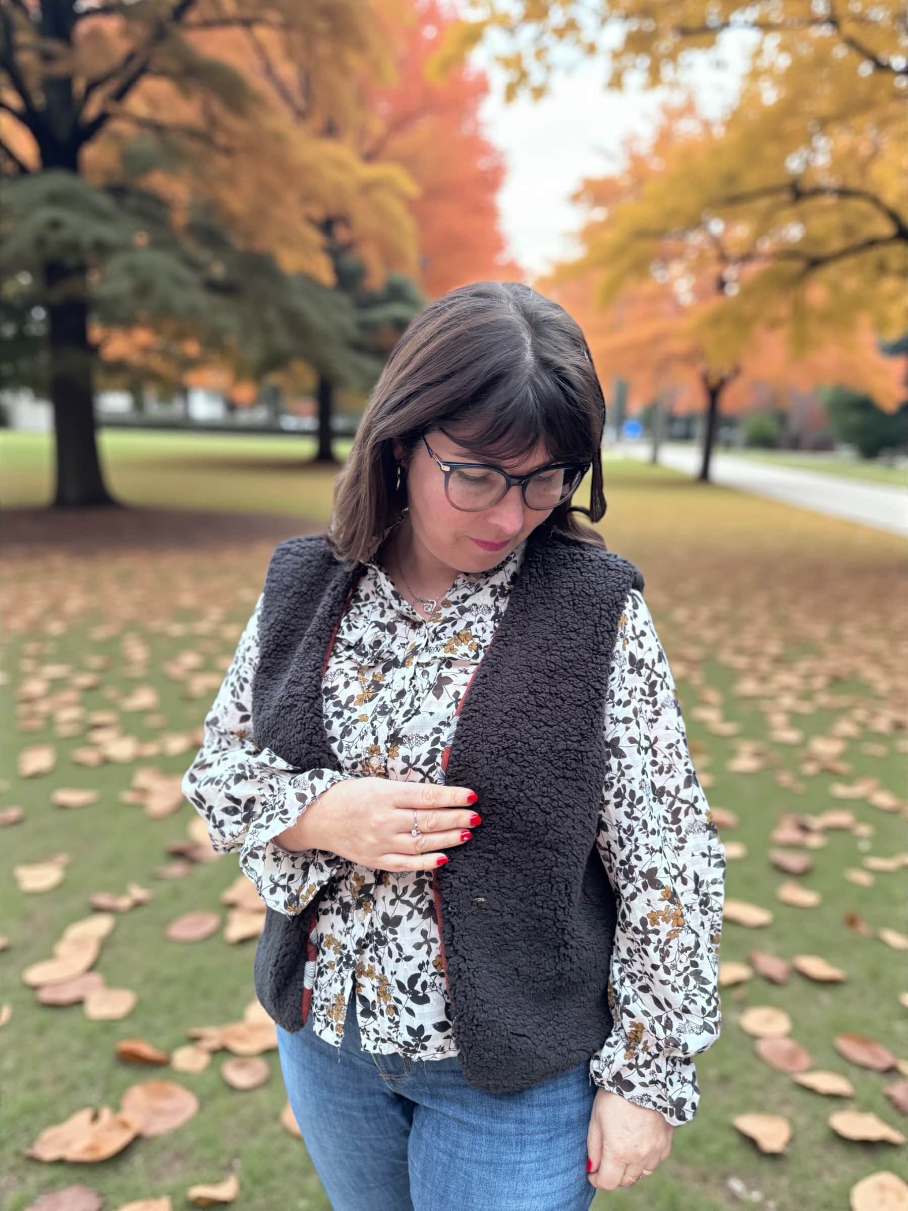 Femme portant un gilet de berger sans manches en sherpa noir par-dessus une blouse fleurie, en extérieur dans un parc avec des feuilles d’automne au sol, regard baissé.