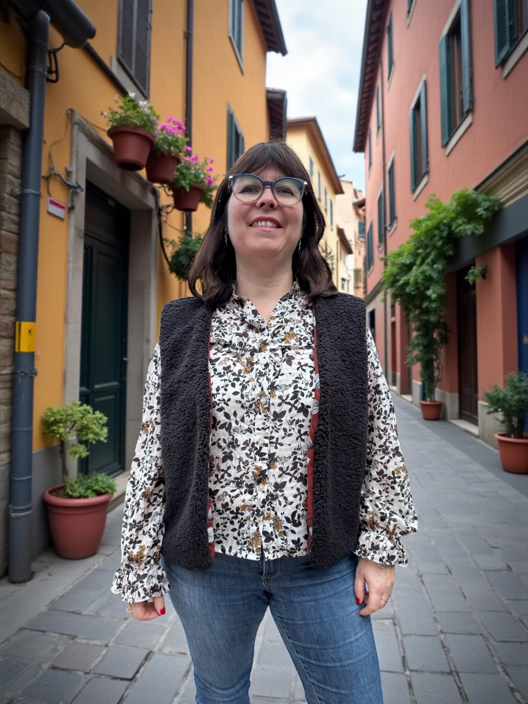 Femme souriante portant un gilet de berger sans manches en sherpa noir, par-dessus une blouse fleurie, dans une ruelle colorée avec des plantes en pots.