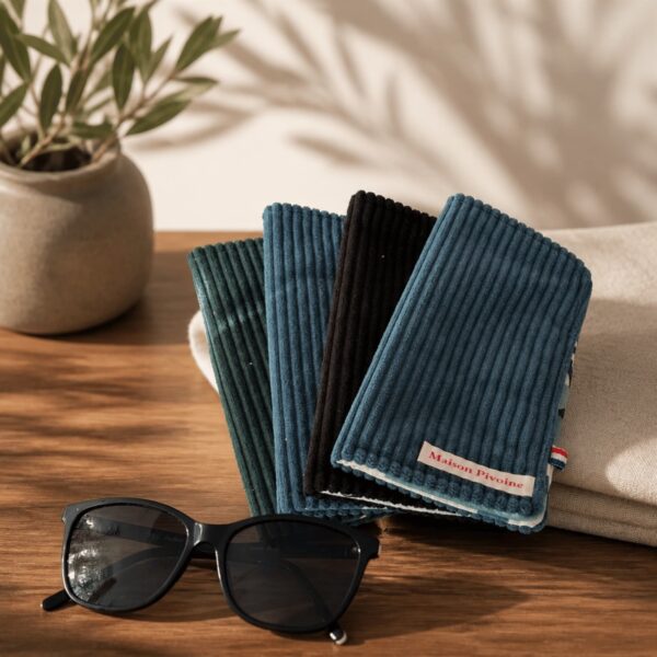 Un empilement de quatre pochettes en velours côtelé (vert forêt, bleu canard, noir, bleu canard) posées sur une table en bois, à côté d'une plante en pot et d'un drap de bain beige. Des ombres portées de feuilles sont projetées sur le mur en arrière-plan. Les lunettes de soleil noires sont posées au premier plan à gauche. Le label "Maison Pivoine" est visible sur la pochette supérieure.